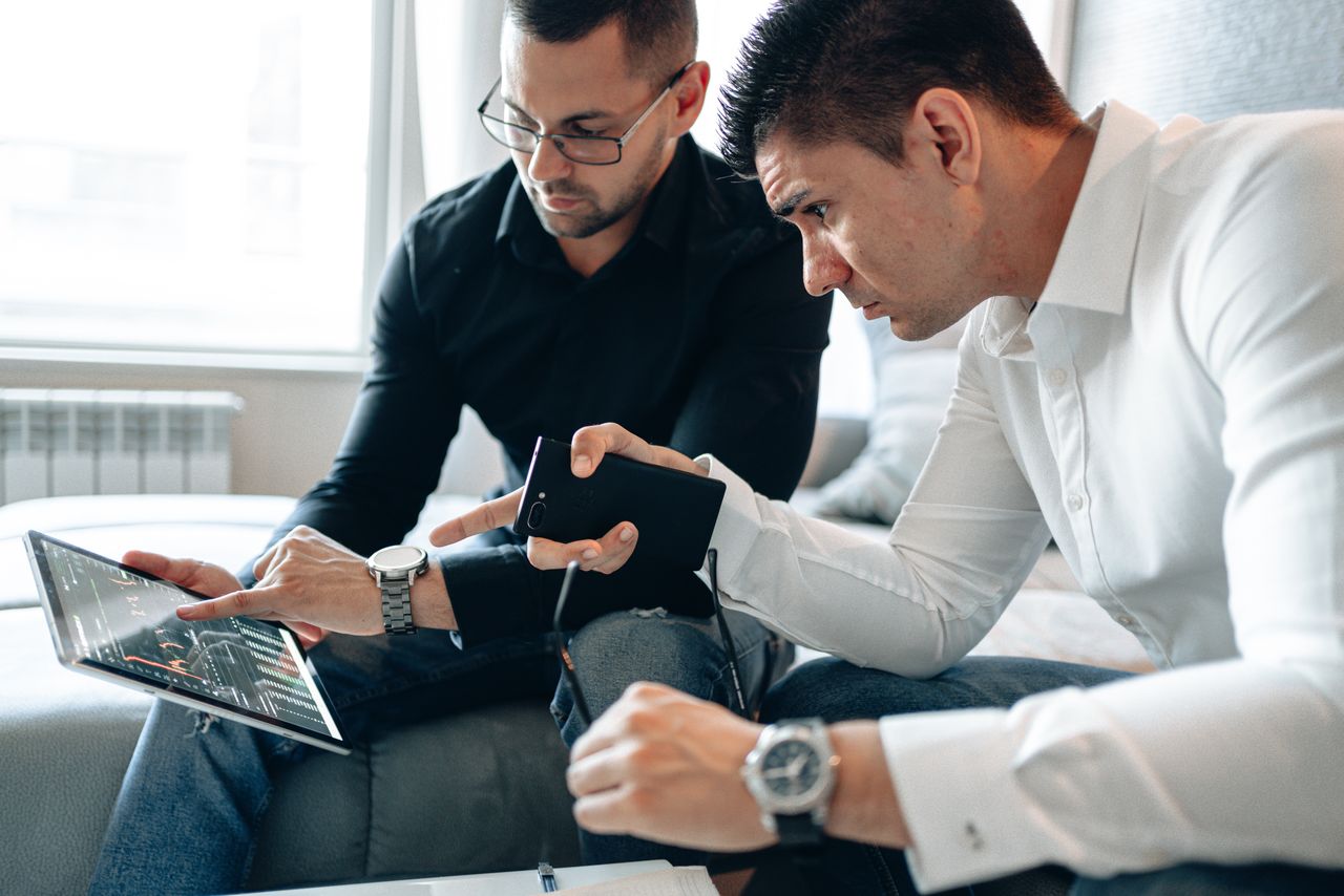 about-01 Two men analyzing financial data on a digital tablet indoors in a professional setting.